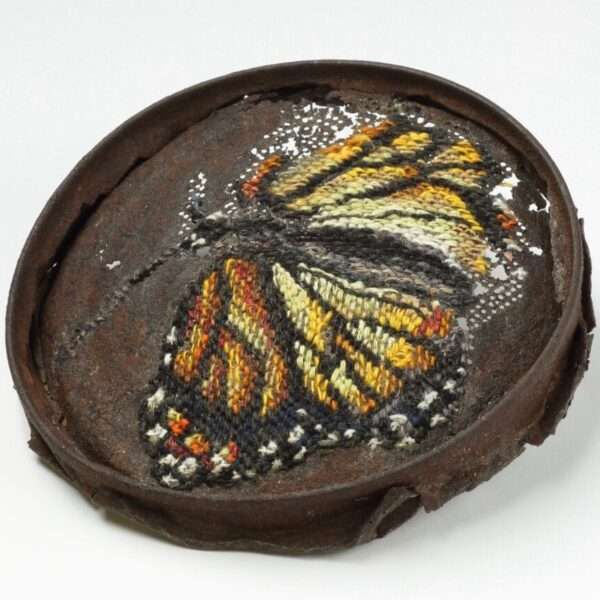 a rusted, round piece of metal with a cross-stitched butterfly made through perforated holes