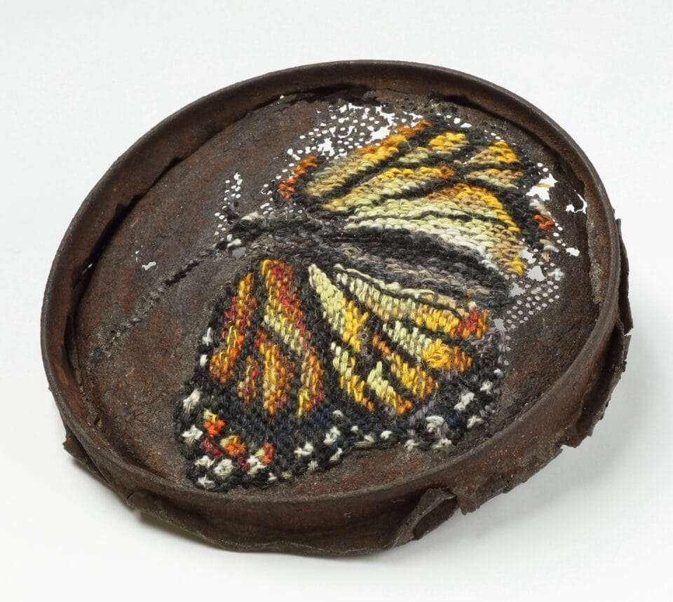 a rusted, round piece of metal with a cross-stitched butterfly made through perforated holes