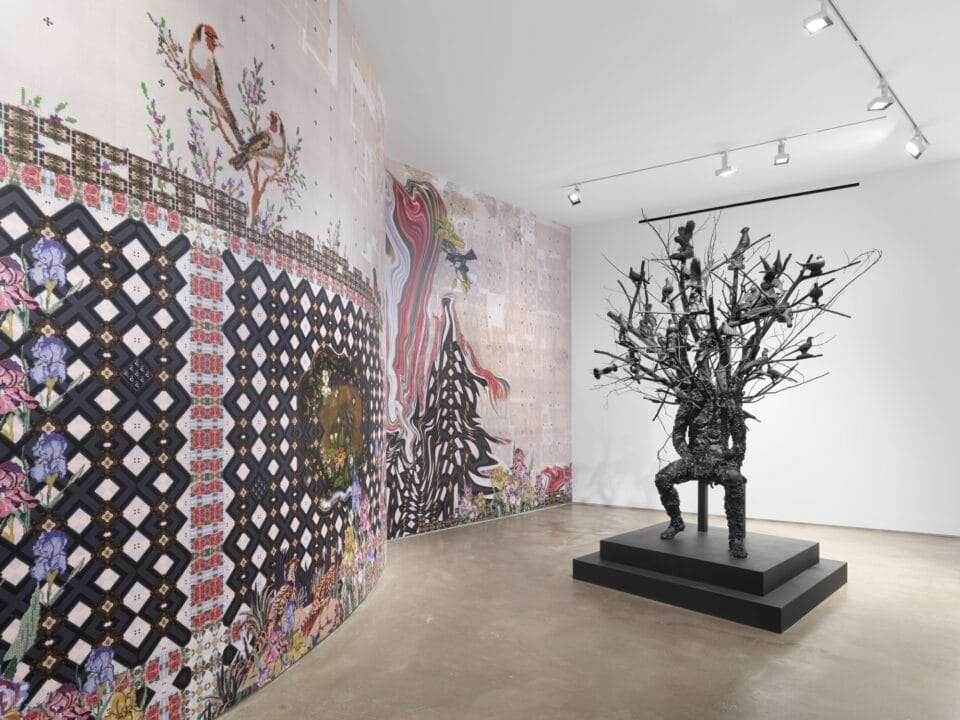 a large-scale bronze figure with branches and leaves for a head sits on a chair next to elaborate wallpaper