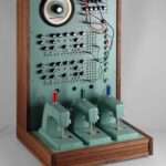 a sculpture resembling a retro computer or device in a teal hue with wood on the sides, with dials and wires and a row of three sewing machines on the base