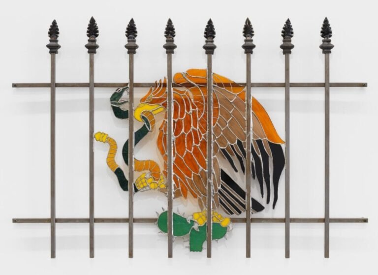 a wall sculpture of stained glass depicting an eagle with a snake in its mouth similar to the Mexican flag emblem, installed behind a fence