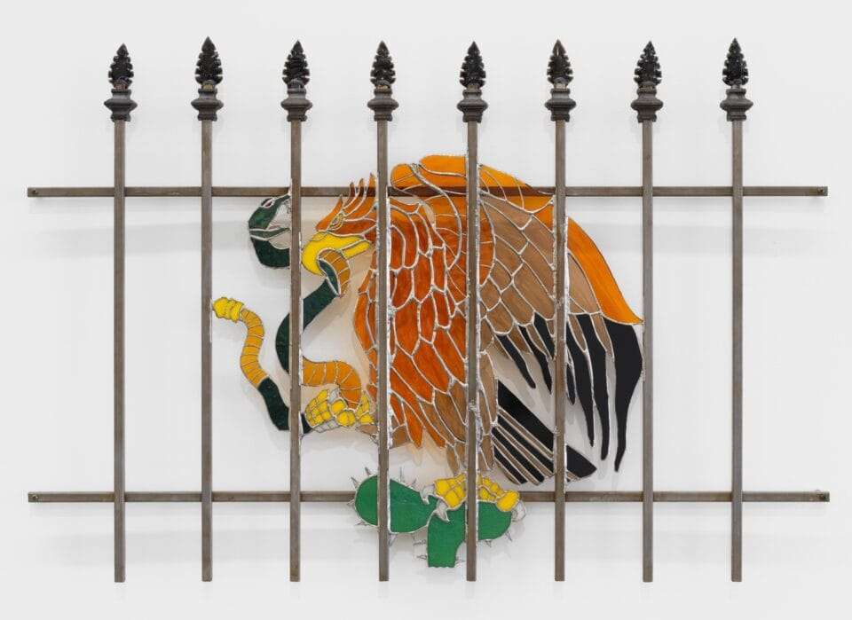 a wall sculpture of stained glass depicting an eagle with a snake in its mouth similar to the Mexican flag emblem, installed behind a fence