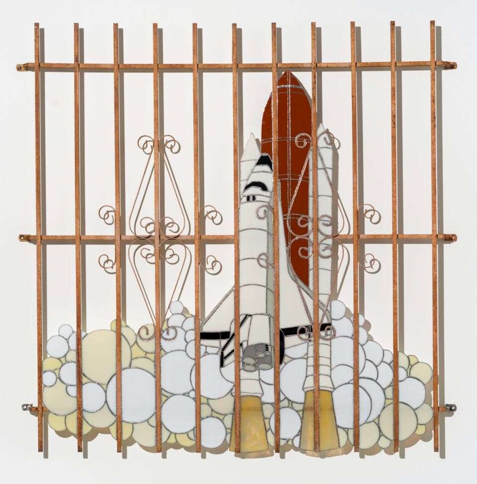 a wall sculpture of stained glass depicting a space shuttle launch, installed behind a fence