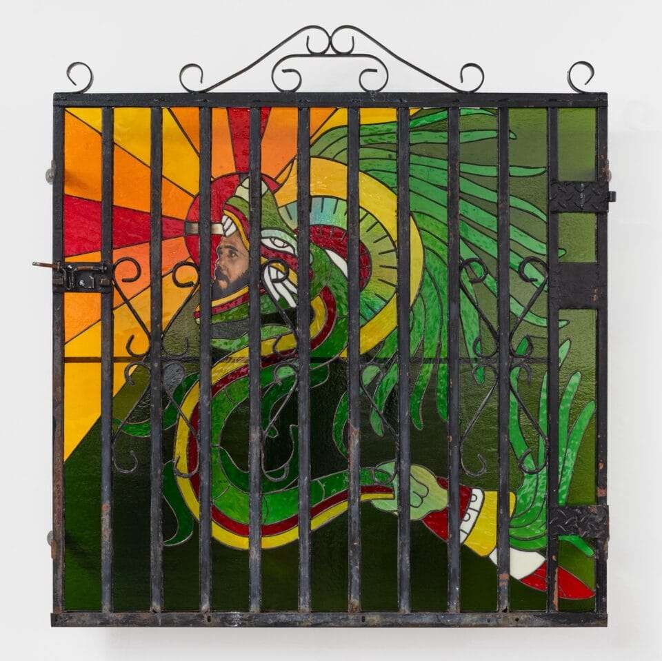 a wall sculpture of stained glass depicting a green dragon with a man's face in its mouth, installed behind a fence