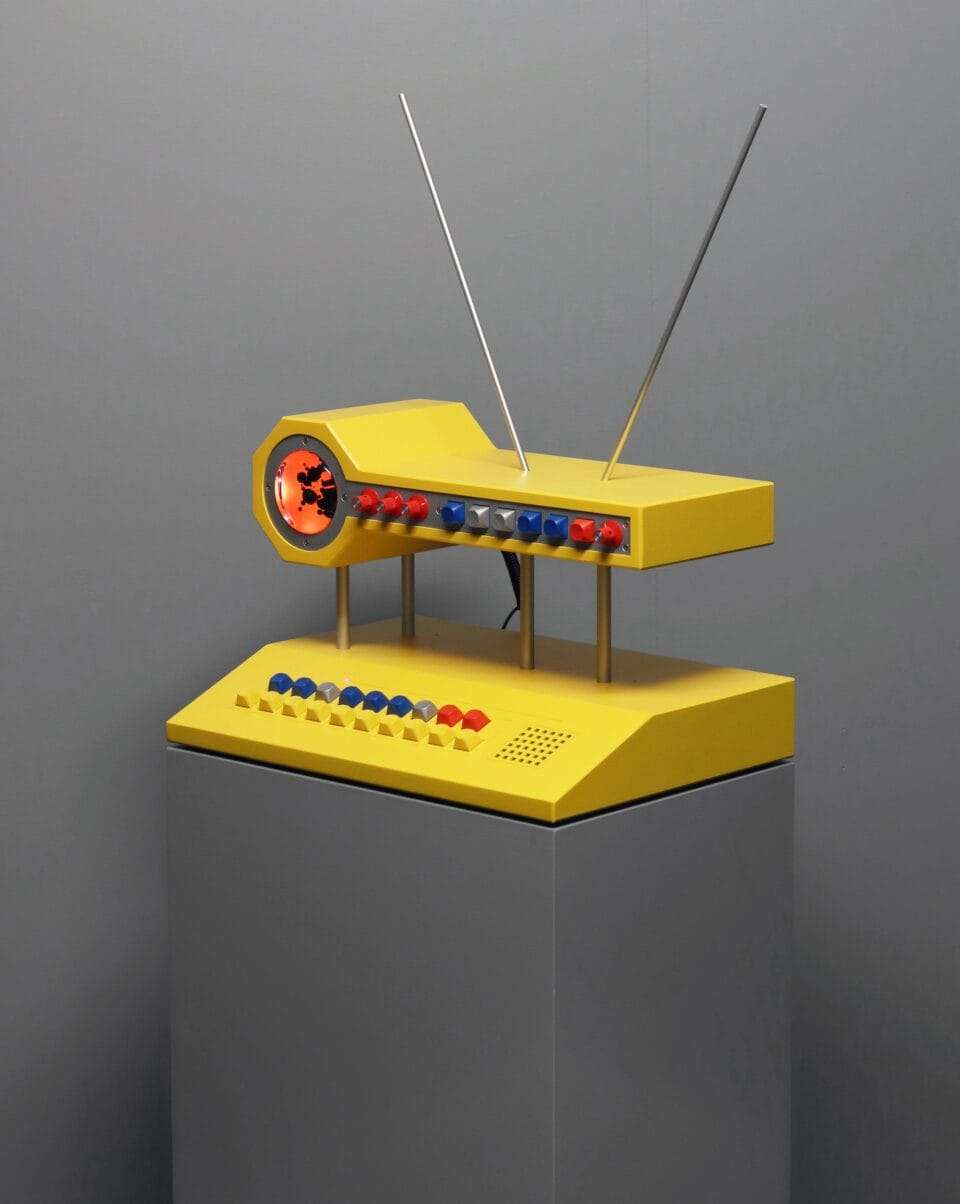 a sculpture resembling a retro computer or device in bright yellow with illuminated orange dials and black and white buttons