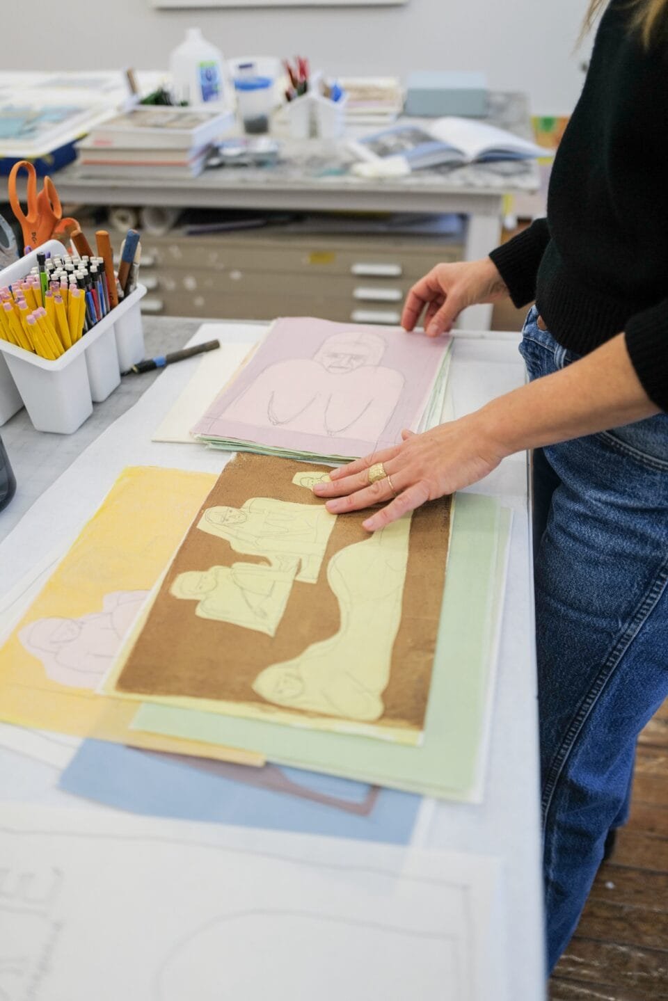 a tabletop in artist Ruby Sky Stiler's studio with sketches for new paintings, shown as the artist picks them up and displays a couple