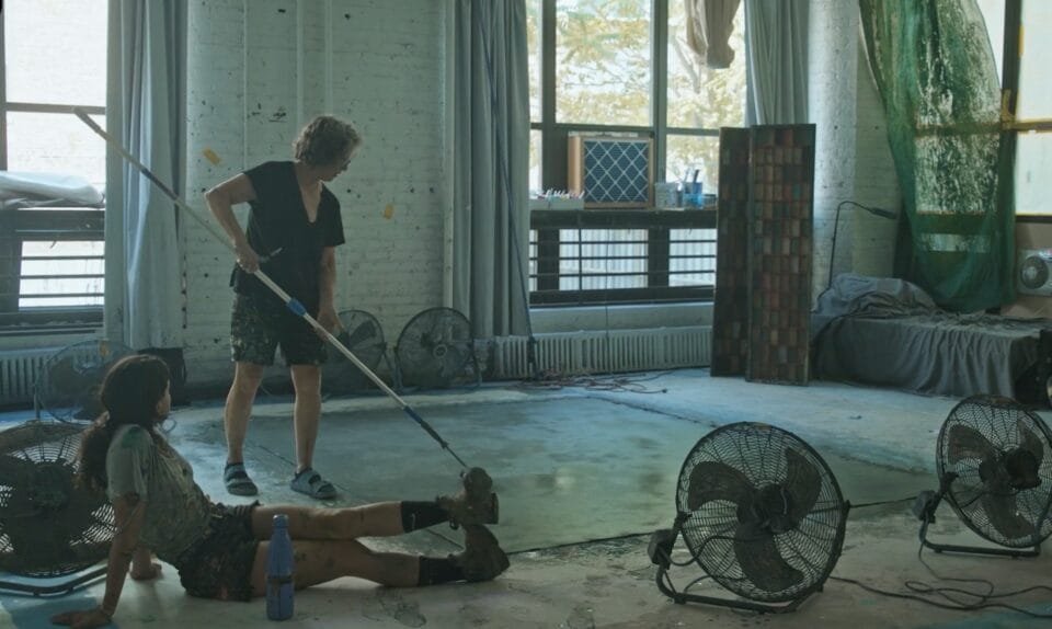 a younger woman sits on the floor while an older woman paints a canvas with a roller