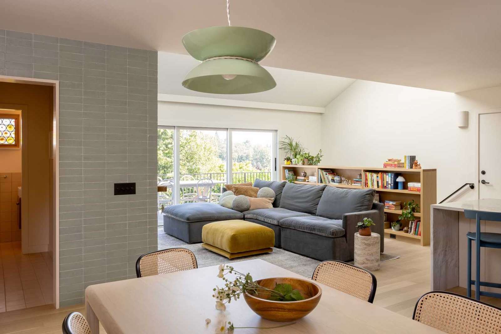 An updated living room includs vaulted ceiling, complemented by a series of skylights, provides a generous, airy feel, filling the space with natural light. A striking green glazed brick fireplace serves as a focal point, extending upward into the skylight and drawing the eye.