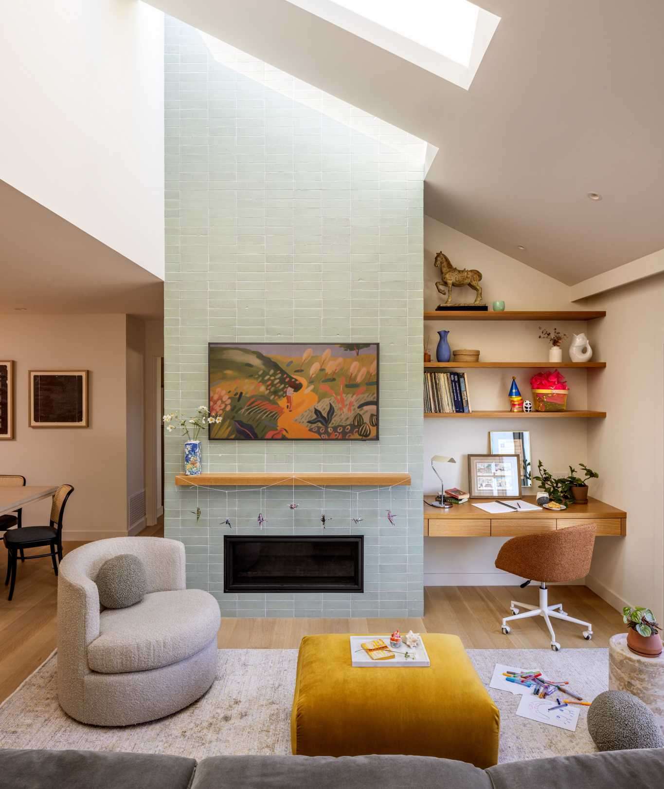 An updated living room includs vaulted ceiling, complemented by a series of skylights, provides a generous, airy feel, filling the space with natural light. A striking green glazed brick fireplace serves as a focal point, extending upward into the skylight and drawing the eye.