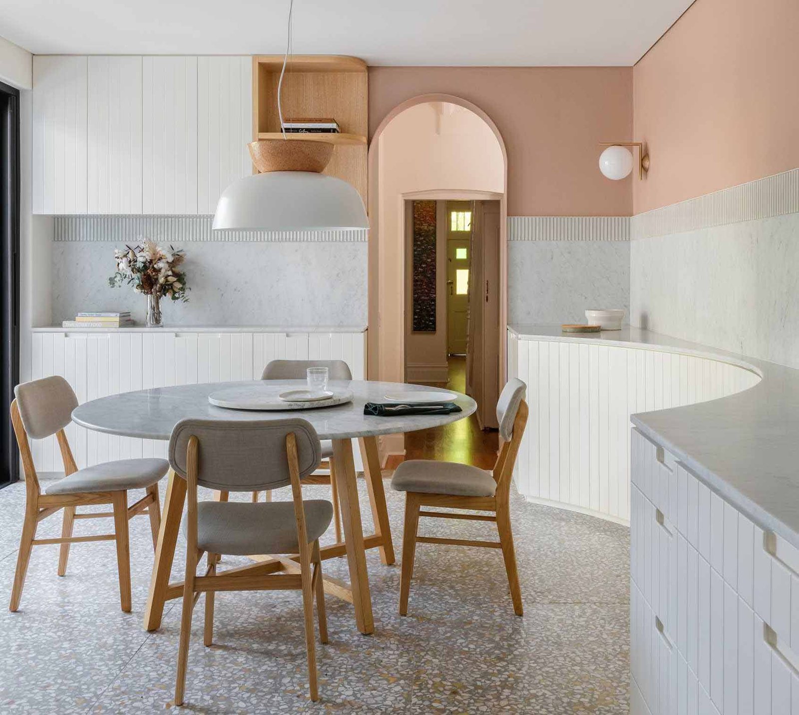 In this modern dining area, earthy peach painted above the tile datum helps to centre and hold the space, while a circular Jardan marble dining table is central to the space, and a curved timber panelled joinery lines the wall.
