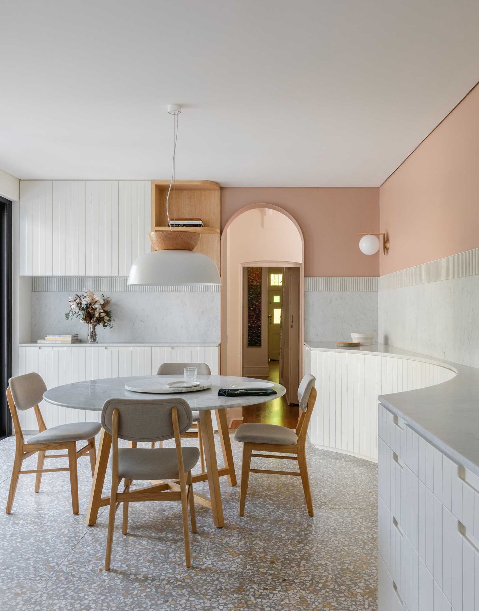 In this modern dining area, earthy peach painted above the tile datum helps to centre and hold the space, while a circular Jardan marble dining table is central to the space, and a curved timber panelled joinery lines the wall.