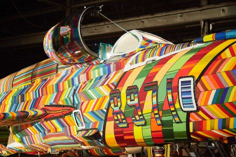 a photograph of a fighter jet coated in colorful beaded patterns, sitting inside a hangar or warehouse