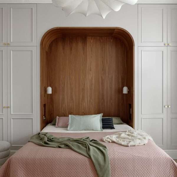 The bedroom blends warmth, softness, and subtle historic detail. A custom walnut niche wraps the bed in a cozy curve, with built-in lights and shelving for quiet functionality. Pale grey wardrobes with brass knobs frame the space, while blush bedding, sage accents, and a sculptural pendant light add layers of texture and calm.