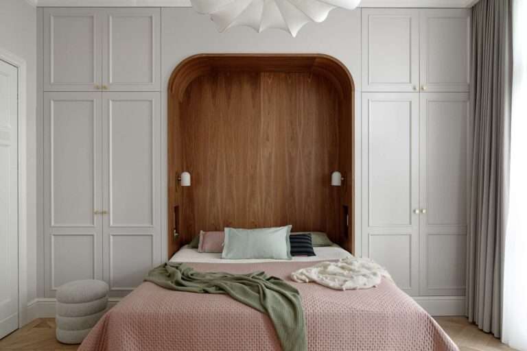 The bedroom blends warmth, softness, and subtle historic detail. A custom walnut niche wraps the bed in a cozy curve, with built-in lights and shelving for quiet functionality. Pale grey wardrobes with brass knobs frame the space, while blush bedding, sage accents, and a sculptural pendant light add layers of texture and calm.