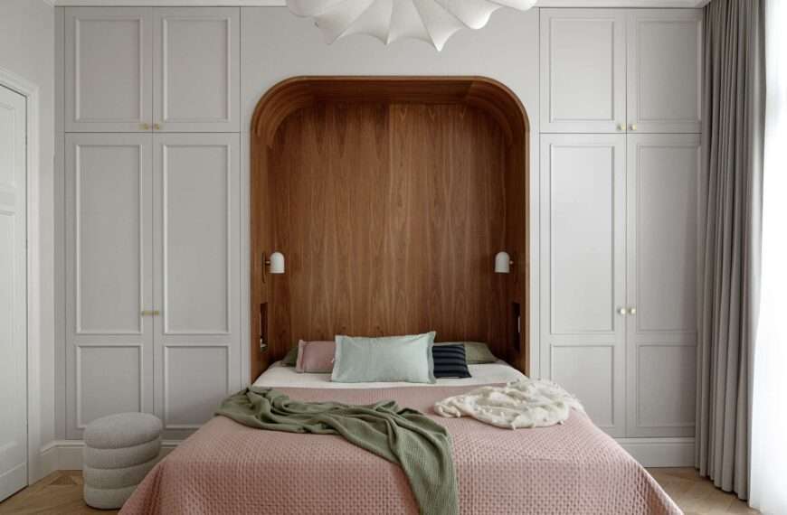 The bedroom blends warmth, softness, and subtle historic detail. A custom walnut niche wraps the bed in a cozy curve, with built-in lights and shelving for quiet functionality. Pale grey wardrobes with brass knobs frame the space, while blush bedding, sage accents, and a sculptural pendant light add layers of texture and calm.