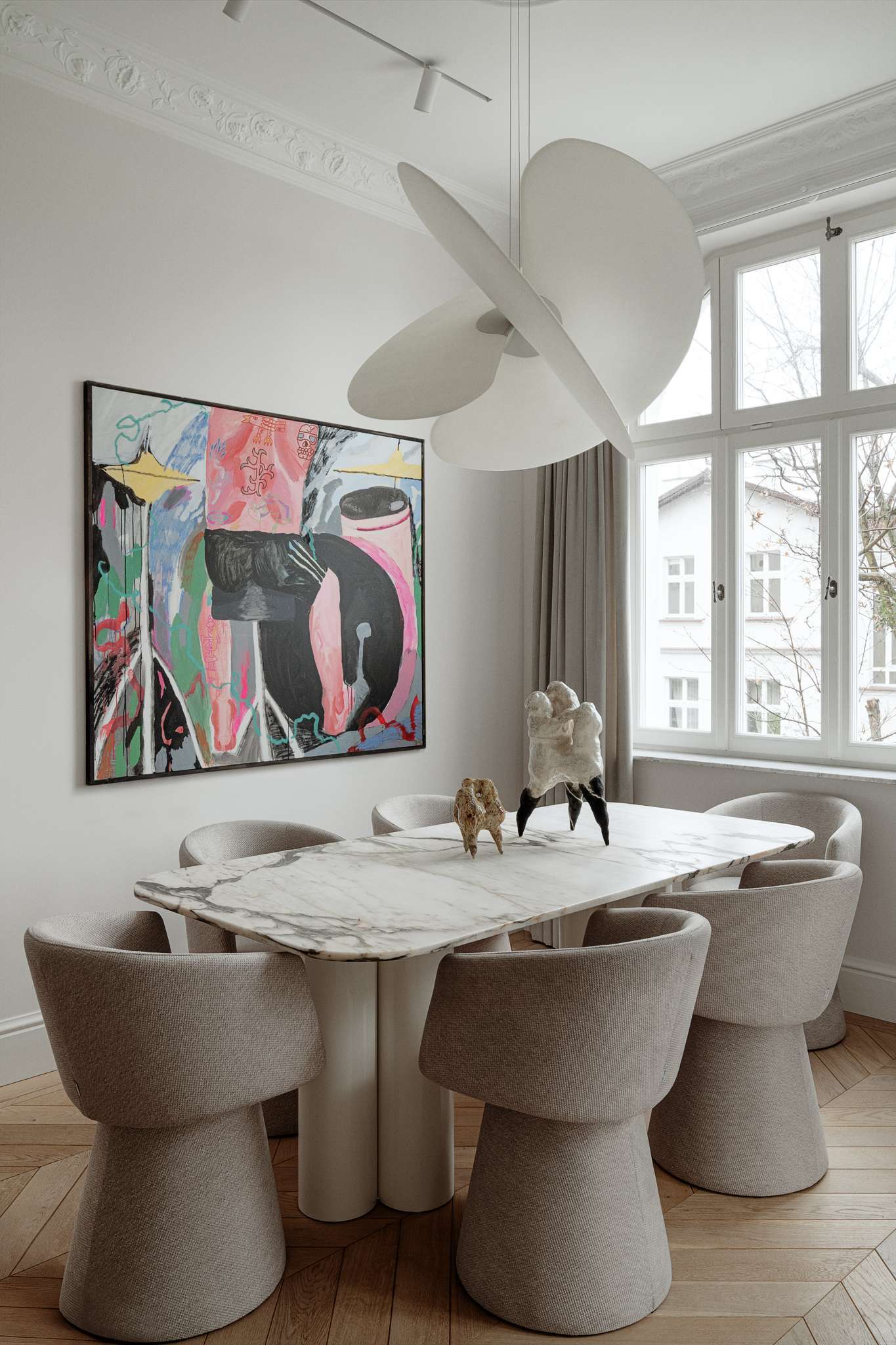 In this dining room, sculptural forms and expressive art define the atmosphere. A veined marble table with softly rounded edges anchors the space, surrounded by textured chairs that echo the home’s gentle curves. Above, a dramatic pendant light introduces a sense of motion and softness, while a large, abstract painting by Maciej Salomon adds bold contrast to the otherwise muted palette.