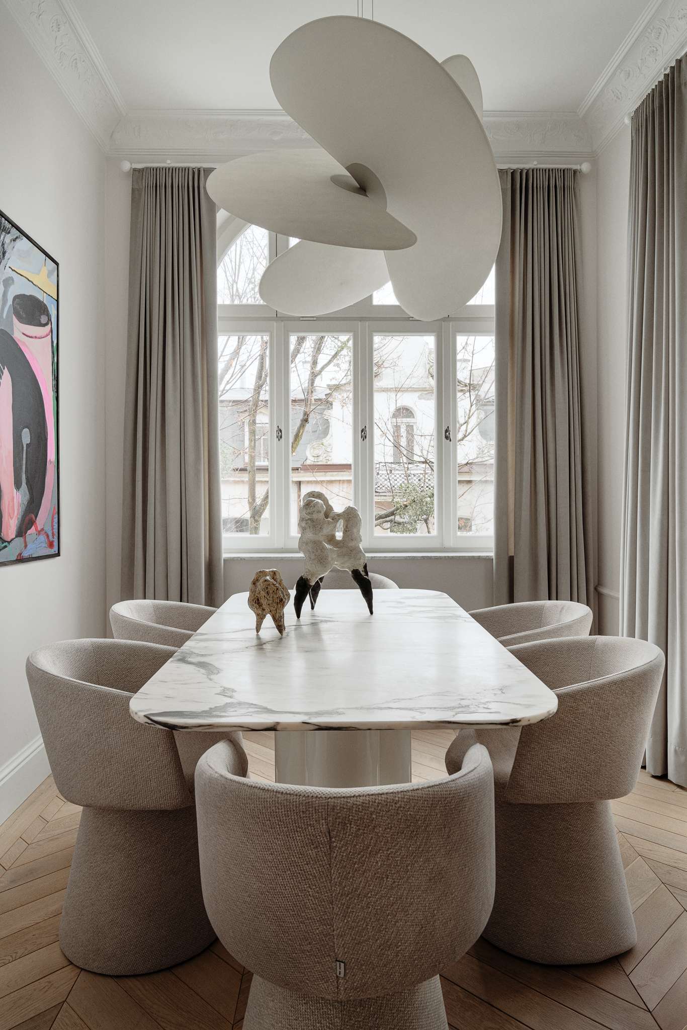 In this dining room, sculptural forms and expressive art define the atmosphere. A veined marble table with softly rounded edges anchors the space, surrounded by textured chairs that echo the home’s gentle curves. Above, a dramatic pendant light introduces a sense of motion and softness, while a large, abstract painting by Maciej Salomon adds bold contrast to the otherwise muted palette.