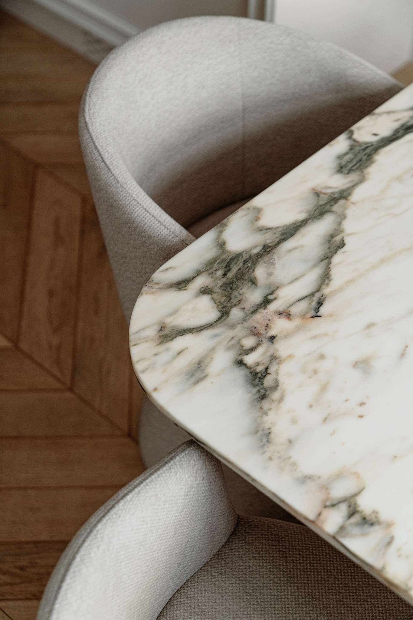 In this dining room, sculptural forms and expressive art define the atmosphere. A veined marble table with softly rounded edges anchors the space, surrounded by textured chairs that echo the home’s gentle curves. Above, a dramatic pendant light introduces a sense of motion and softness, while a large, abstract painting by Maciej Salomon adds bold contrast to the otherwise muted palette.