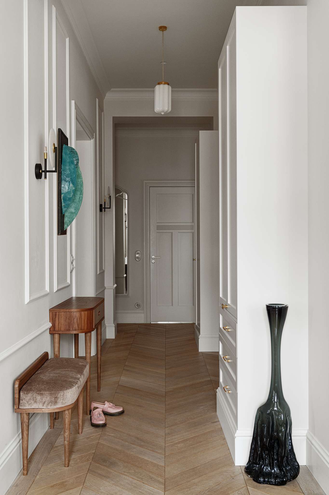 The entryway sets a refined tone with its soft palette, custom paneling, and sculptural details. A slim walnut table and a velvet-upholstered bench create a welcoming spot to pause, while the chevron floor guides the eye through the hallway.