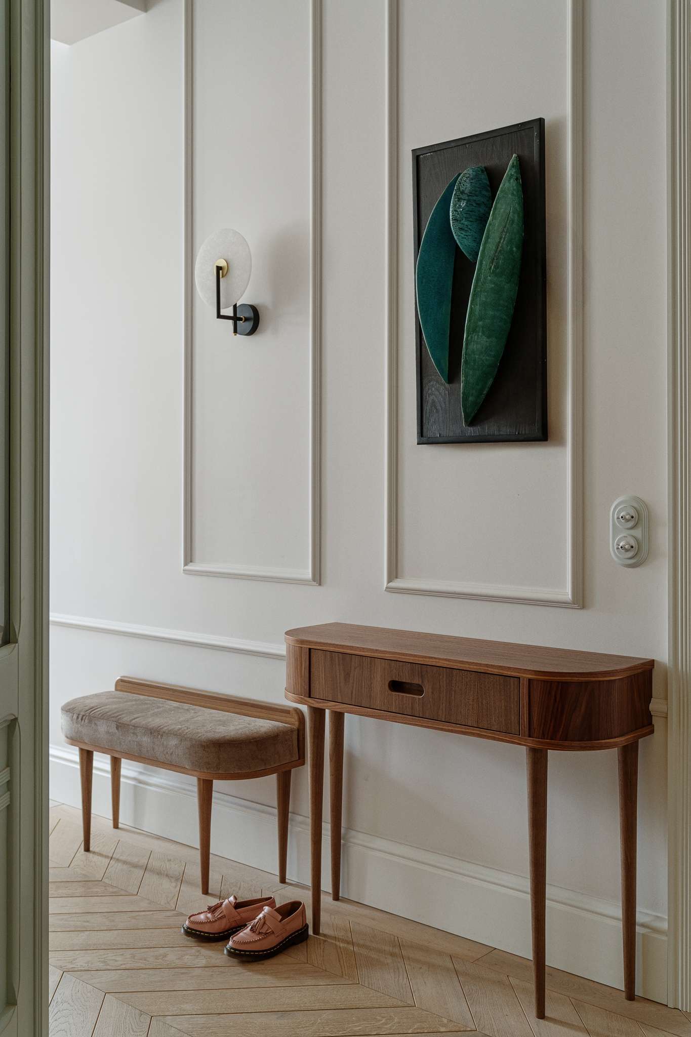 The entryway sets a refined tone with its soft palette, custom paneling, and sculptural details. A slim walnut table and a velvet-upholstered bench create a welcoming spot to pause, while the chevron floor guides the eye through the hallway.
