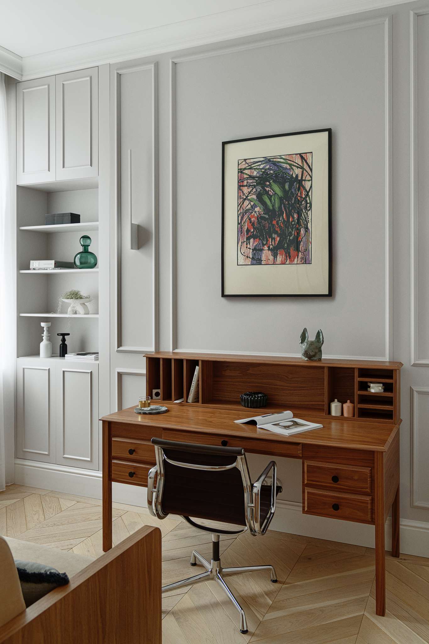 This home office blends functionality with calm, refined design. A wood writing desk brings warmth and structure to the space, its organized cubbies and drawers paired with a modern, chrome-framed chair. Wall panelling and soft grey tones create a subtle architectural backdrop, while built-in shelving offers a curated display of books, ceramics, and sculptural accents.