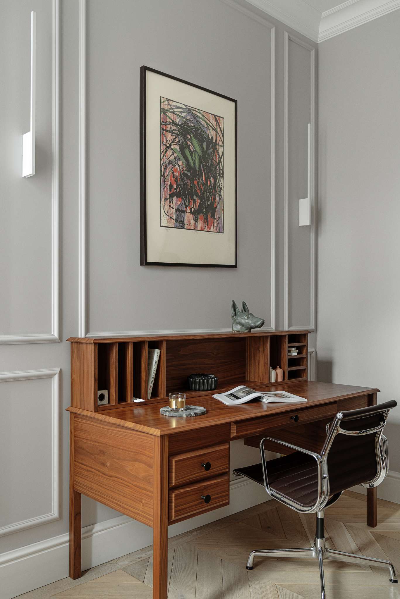This home office blends functionality with calm, refined design. A wood writing desk brings warmth and structure to the space, its organized cubbies and drawers paired with a modern, chrome-framed chair. Wall panelling and soft grey tones create a subtle architectural backdrop, while built-in shelving offers a curated display of books, ceramics, and sculptural accents.