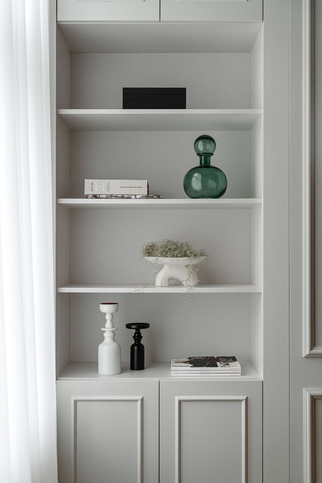 This home office blends functionality with calm, refined design. A wood writing desk brings warmth and structure to the space, its organized cubbies and drawers paired with a modern, chrome-framed chair. Wall panelling and soft grey tones create a subtle architectural backdrop, while built-in shelving offers a curated display of books, ceramics, and sculptural accents.