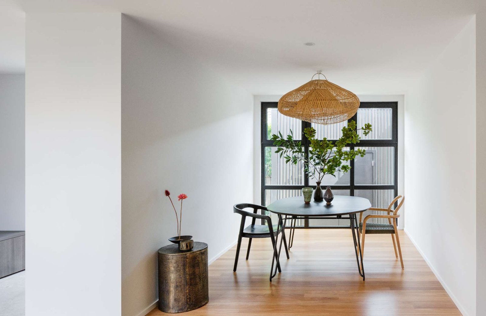 The dining area is calm and restrained, defined by clean lines, soft light and a refined palette. A round table sits beneath a woven pendant, flanked by sculptural chairs in mismatched finishes that add quiet character without disrupting harmony.