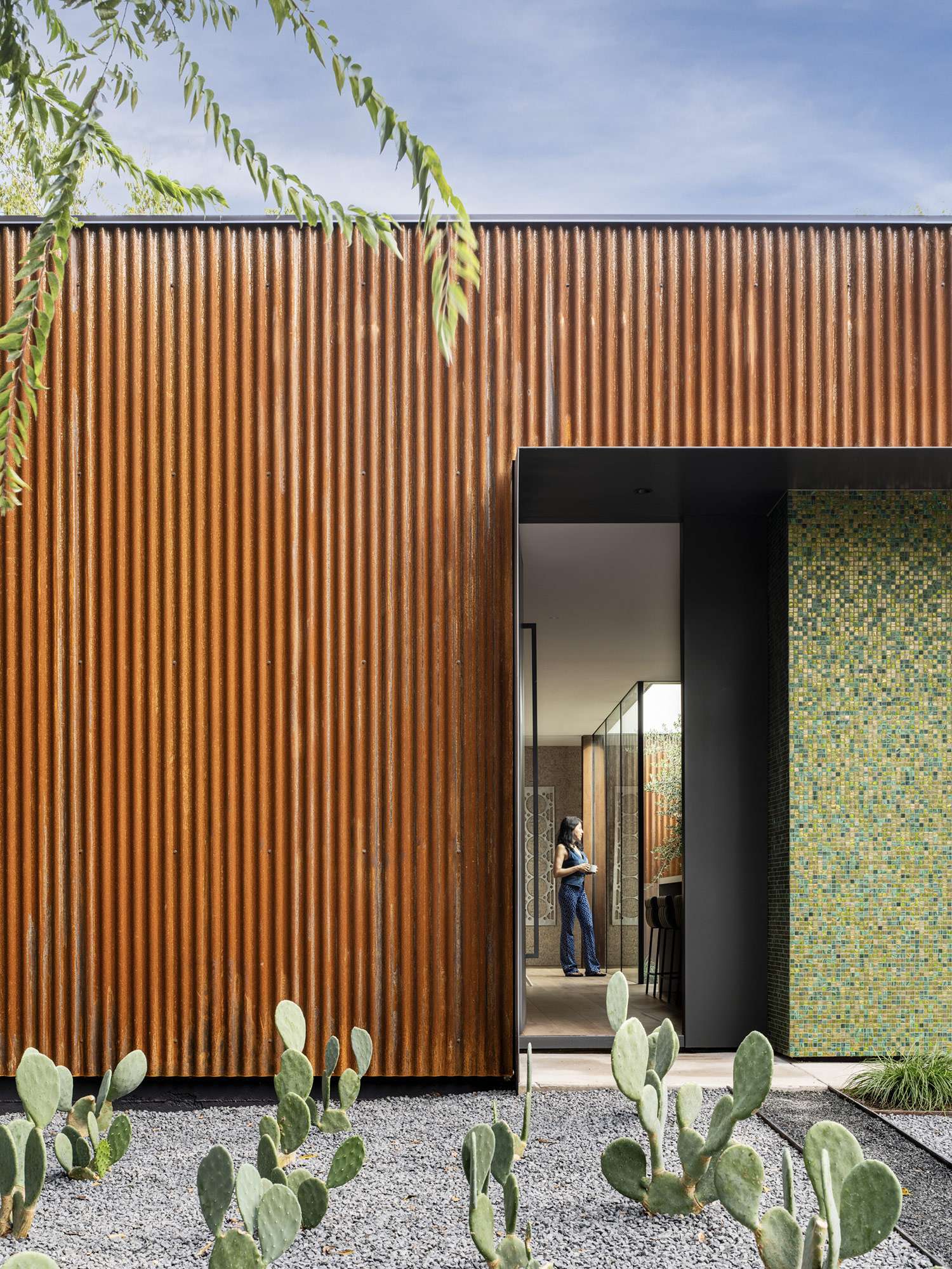 A bold mix of industrial and refined materials, this modern home pairs rusted corrugated steel with a shimmering mosaic wall for a striking first impression. The narrow window offers a glimpse into the tranquil, glass-lined interior.