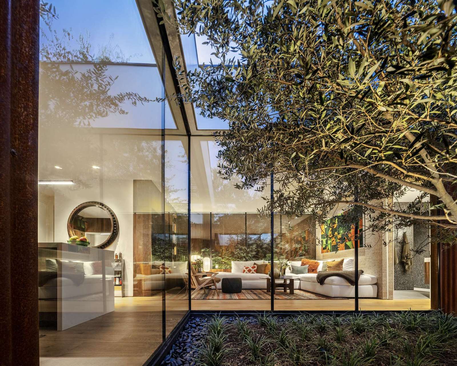 This serene courtyard anchors the home with a sculptural olive tree framed by floor-to-ceiling glass. Natural light floods the kitchen and living spaces while rusted steel cladding brings warmth and texture to the modern design.