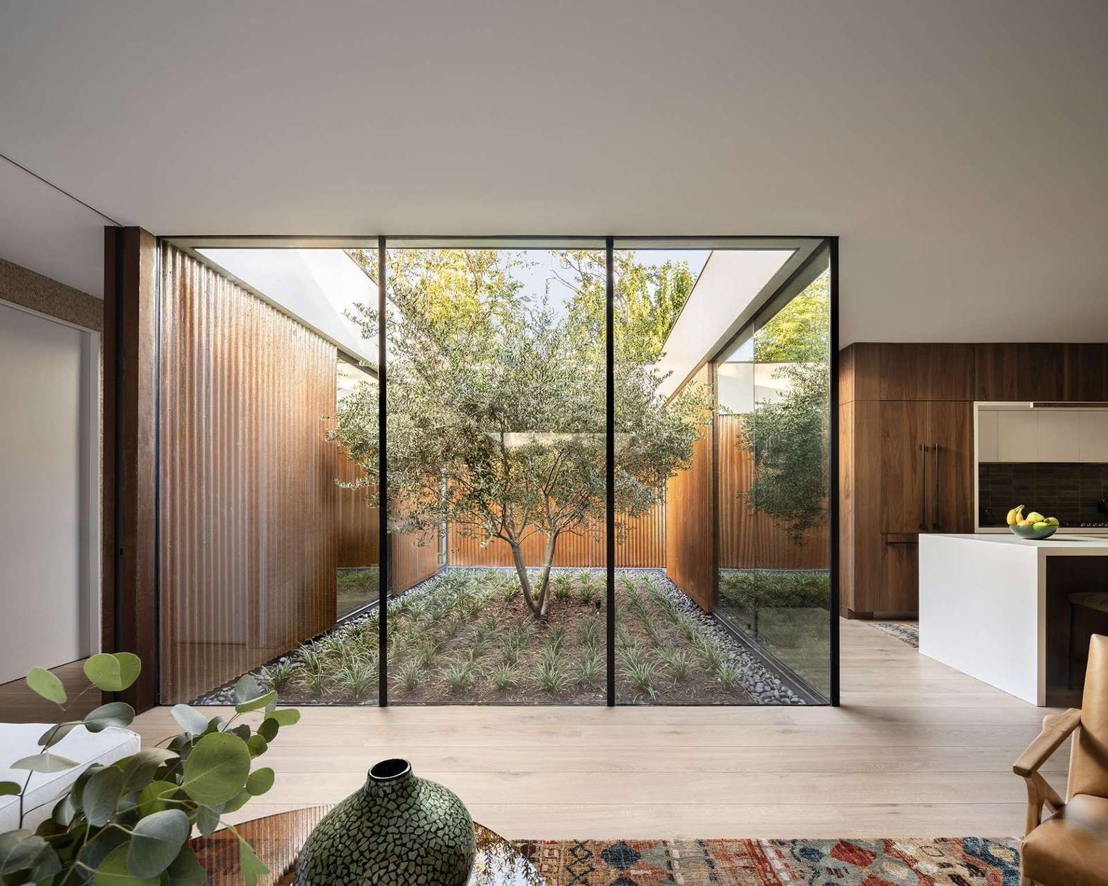 This serene courtyard anchors the home with a sculptural olive tree framed by floor-to-ceiling glass. Natural light floods the kitchen and living spaces while rusted steel cladding brings warmth and texture to the modern design.