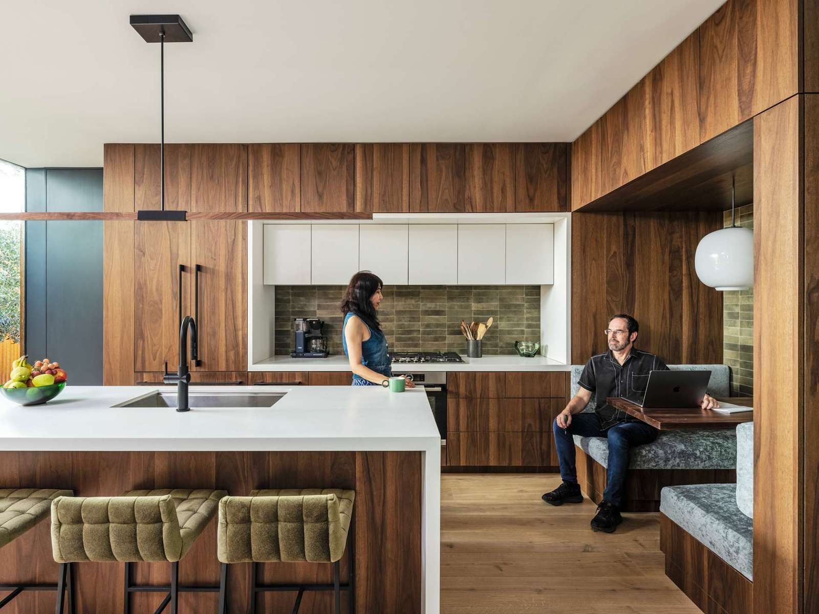 Rich wood cabinetry sets a warm tone in this modern kitchen, where clean white countertops and olive-toned backsplash tiles add depth. A built-in breakfast nook doubles as a cozy workspace, blending form and function beautifully.