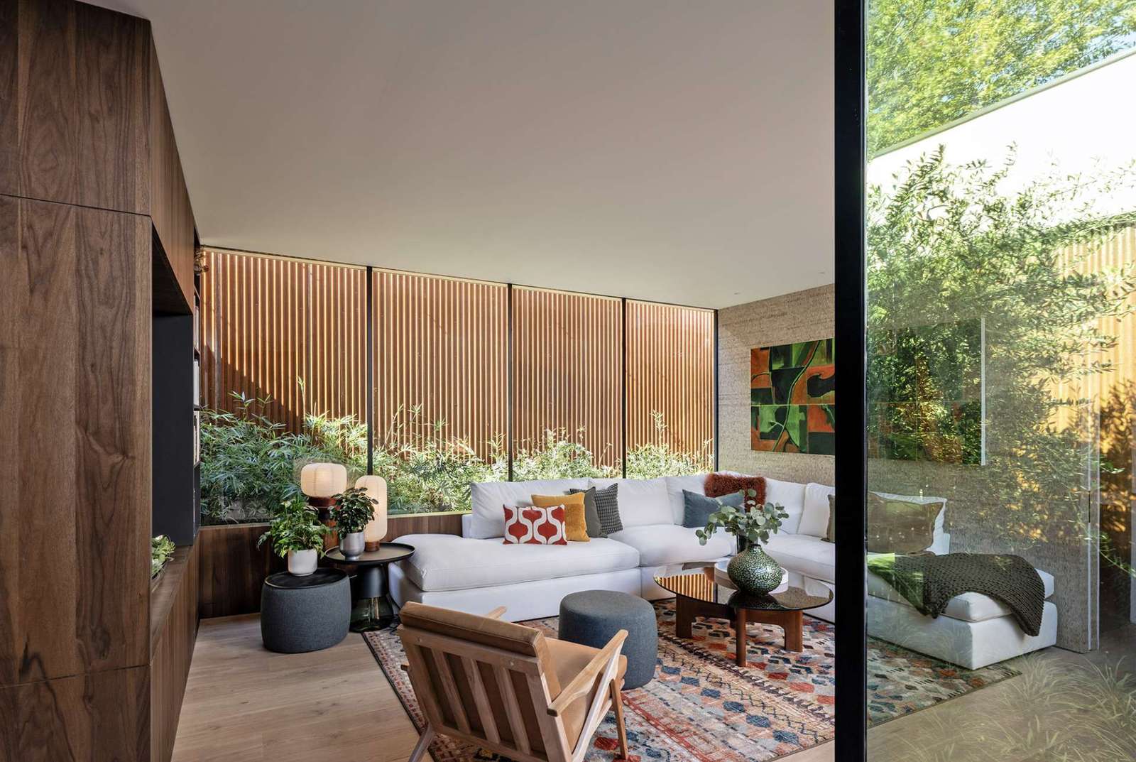This cozy living room blends earthy textures and warm wood tones with expansive glass walls that open to lush bamboo landscaping. A colorful rug and sculptural furnishings add comfort and character to the modern minimalist space.