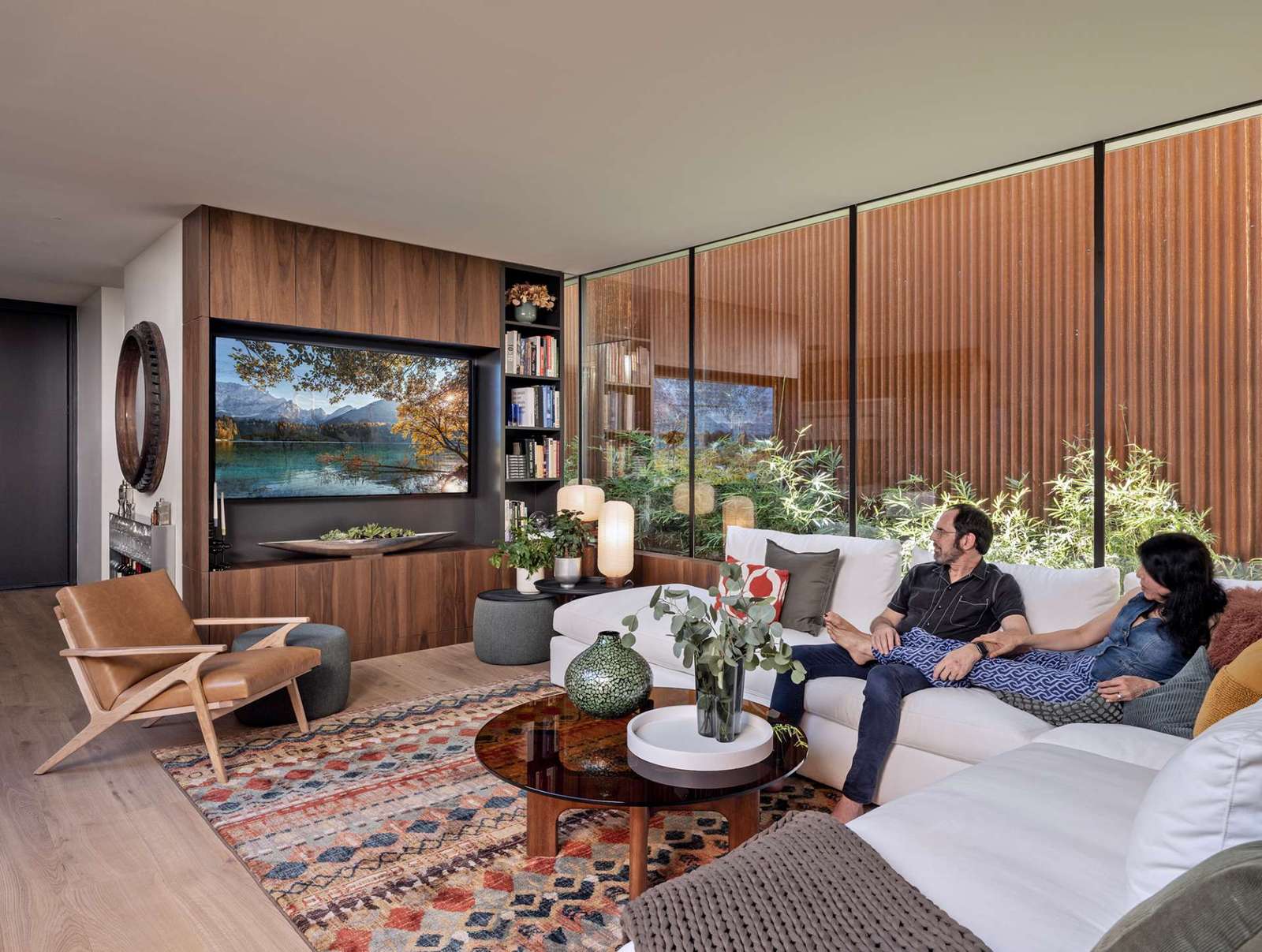 This cozy living room blends earthy textures and warm wood tones with expansive glass walls that open to lush bamboo landscaping. A colorful rug and sculptural furnishings add comfort and character to the modern minimalist space.