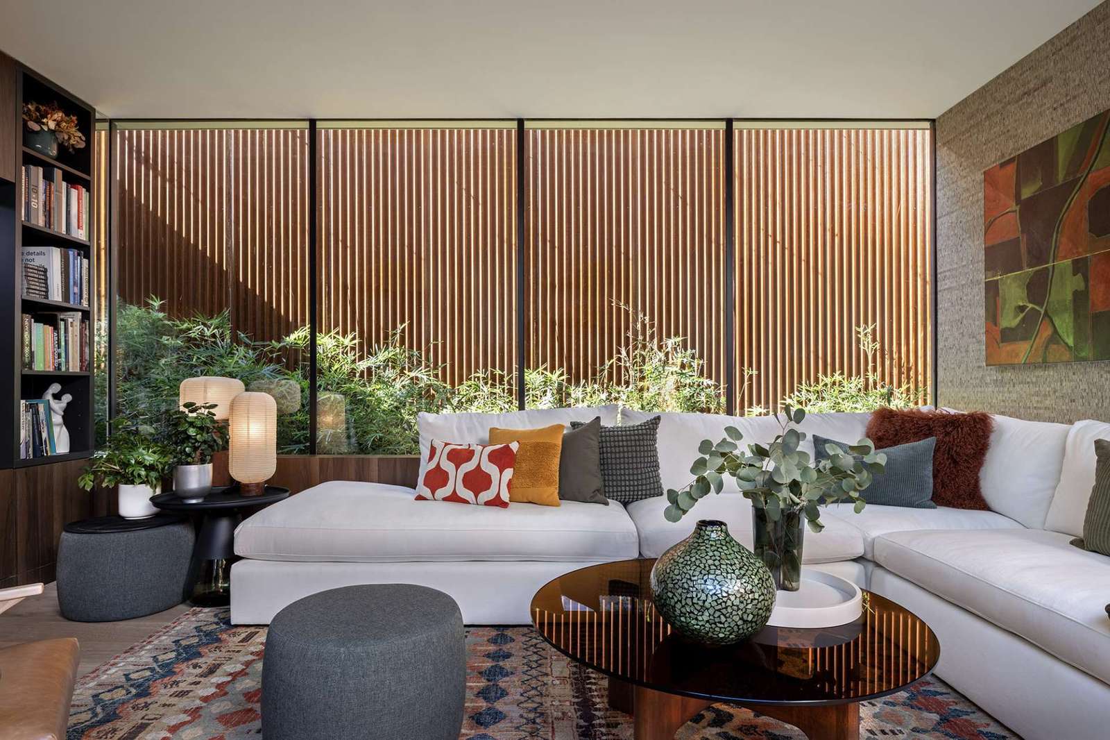 This cozy living room blends earthy textures and warm wood tones with expansive glass walls that open to lush bamboo landscaping. A colorful rug and sculptural furnishings add comfort and character to the modern minimalist space.