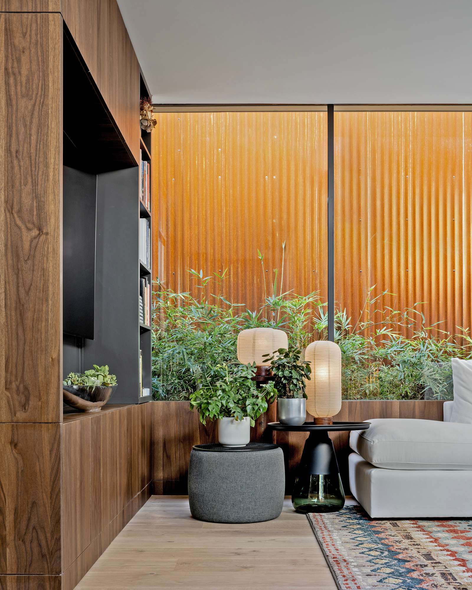 This cozy living room blends earthy textures and warm wood tones with expansive glass walls that open to lush bamboo landscaping. A colorful rug and sculptural furnishings add comfort and character to the modern minimalist space.