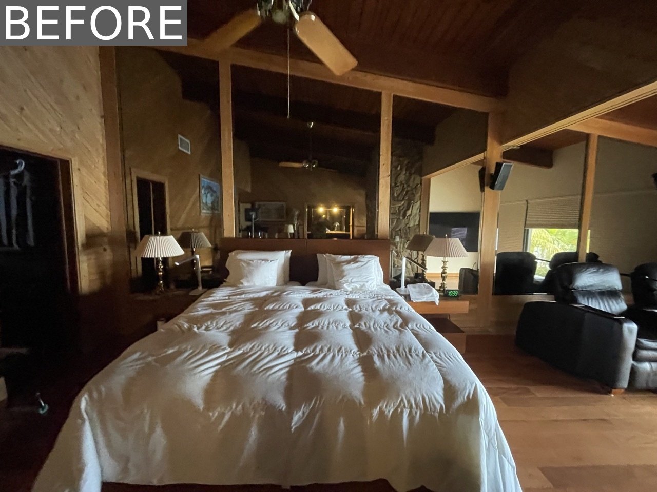 BEFORE - The original primary bedroom felt dark and dated, with a full mirrored wall, a heavy stone accent, and a  pair of oversized black leather recliners focused on a TV.