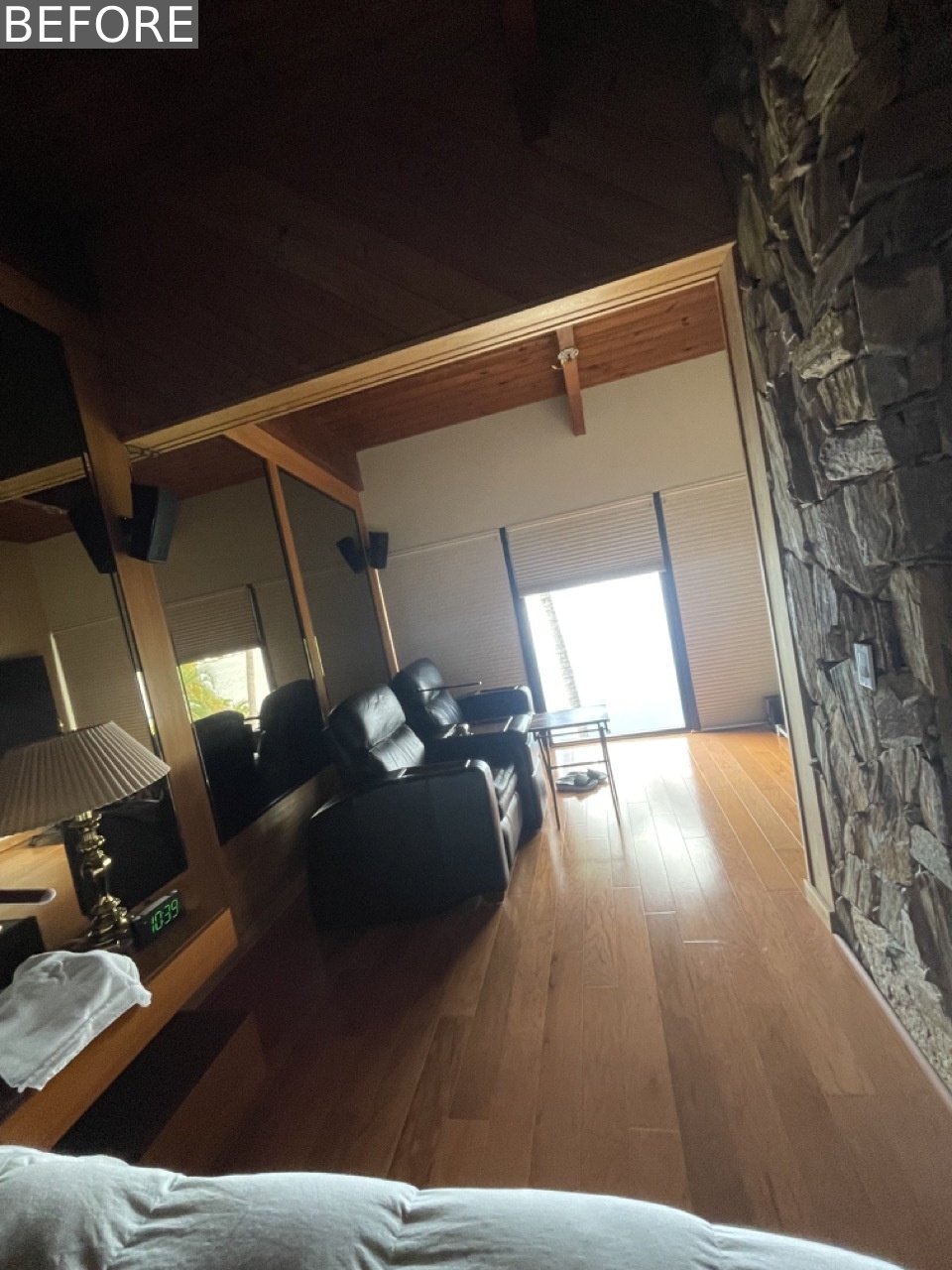 BEFORE - The original primary bedroom felt dark and dated, with a full mirrored wall, a heavy stone accent, and a  pair of oversized black leather recliners focused on a TV.