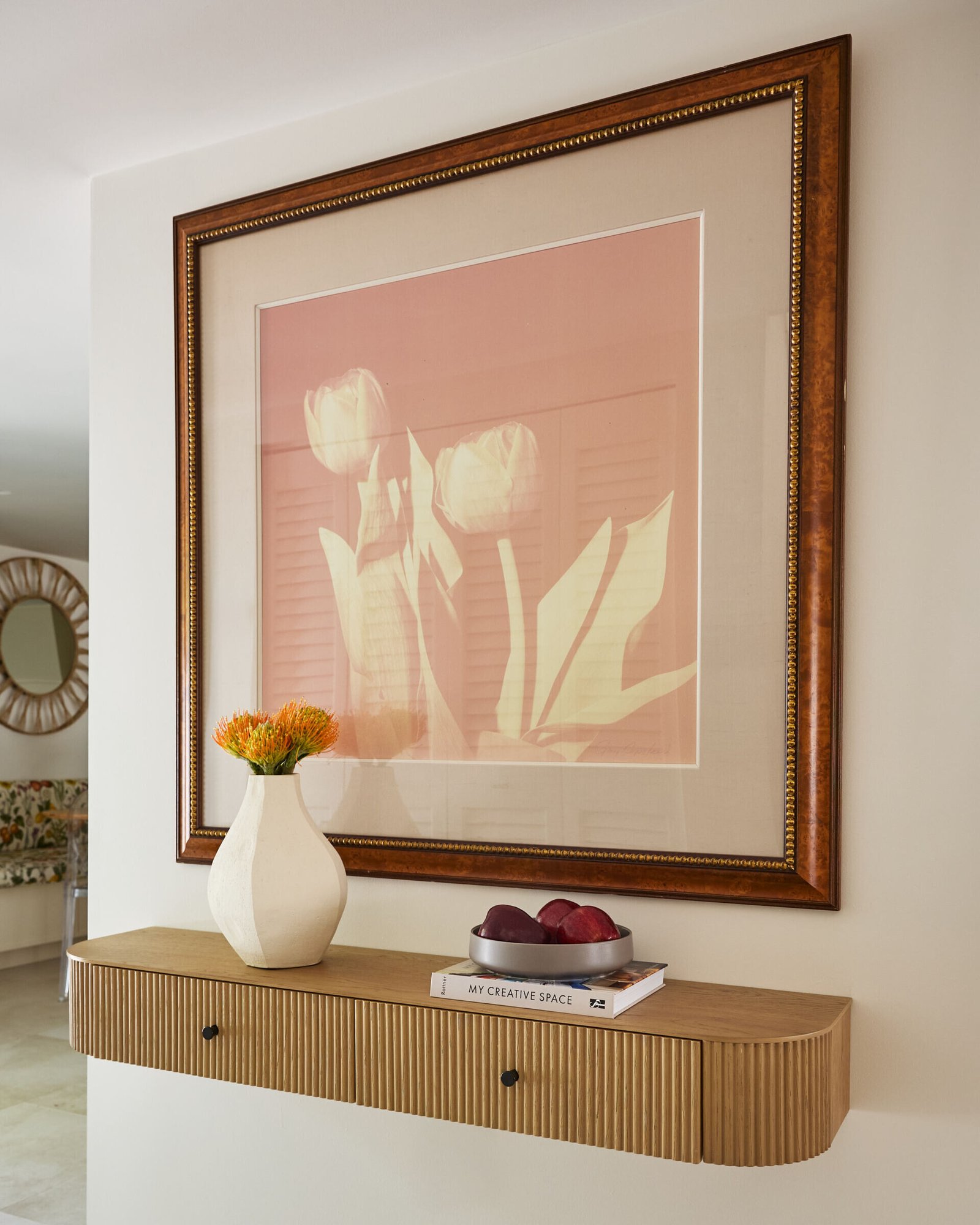 This updated entryway sets the tone immediately. Though not overly grand, the entry is warm and welcoming, offering subtle transitions rather than showy statements. It pairs a fluted floating wood shelf with a soft tulip print and cheerful orange blooms. A small tray of fruit and stacked books add lived-in warmth to the minimalist styling.