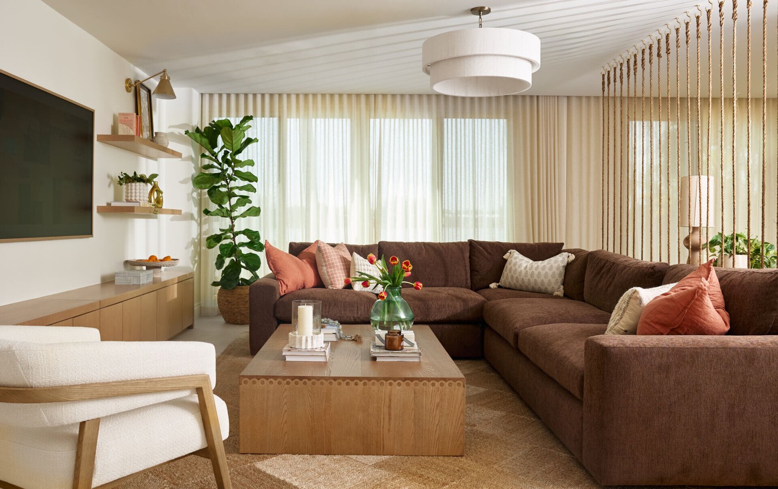 Earthy tones and soft textures define this serene living room. A deep brown sectional, blush accents, and layered neutral rugs create a cozy, light-filled space perfect for everyday comfort.