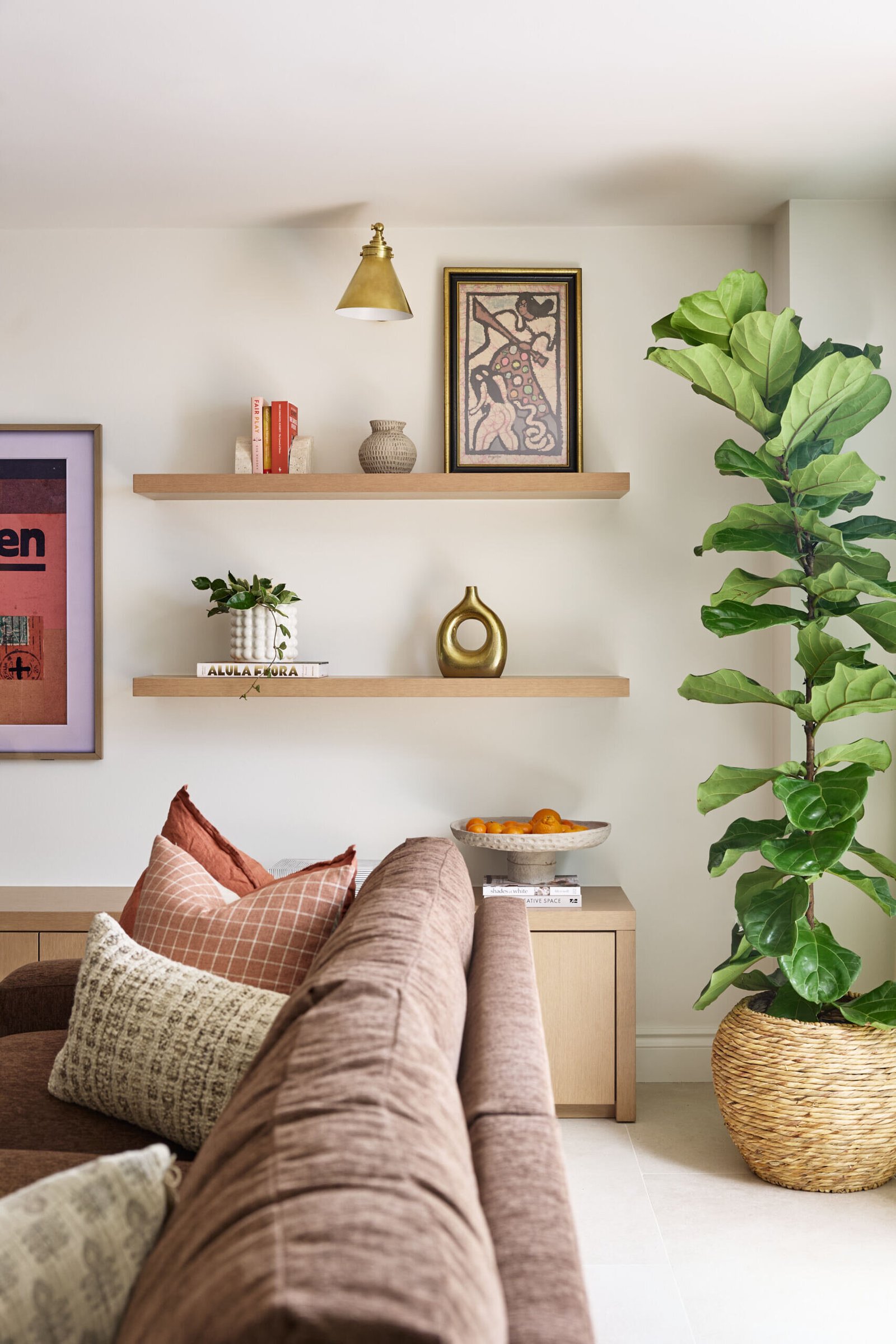 This cozy corner features warm rust pillows, a woven basket with greenery, and curated shelves filled with art and pottery. A thoughtful mix of textures makes it feel lived-in and personal.