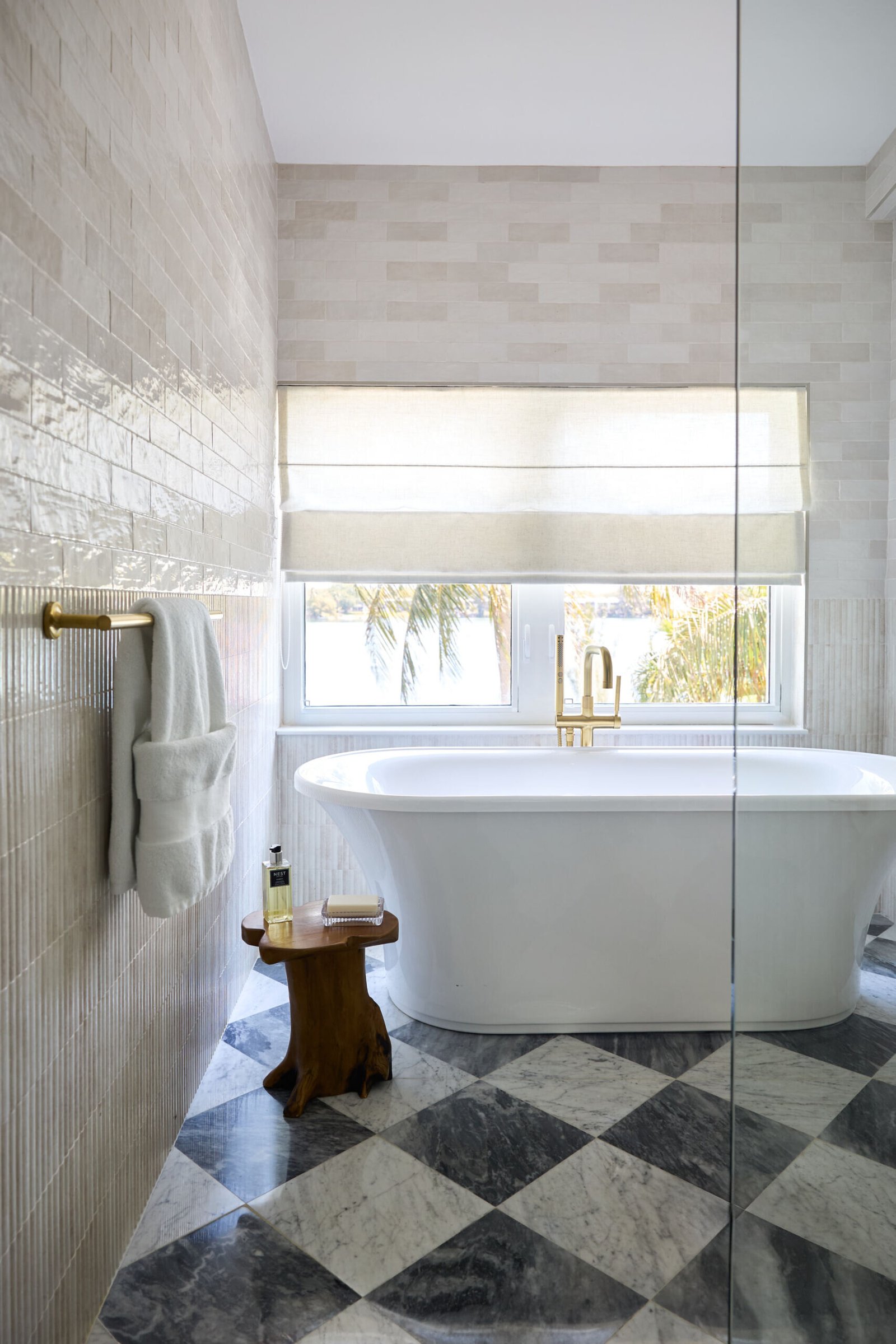 In this primary bath, quiet indulgence is the theme. The checkerboard marble floor adds a timeless graphic punch, offset by creamy wall tiles that bring in softness. A freestanding tub basks in sunlight and lake views, while the custom oak vanity topped with honed marble strikes a clean yet classic tone. Brass fixtures, gold-rimmed mirrors, and alabaster sconces round out the palette in glowing harmony.