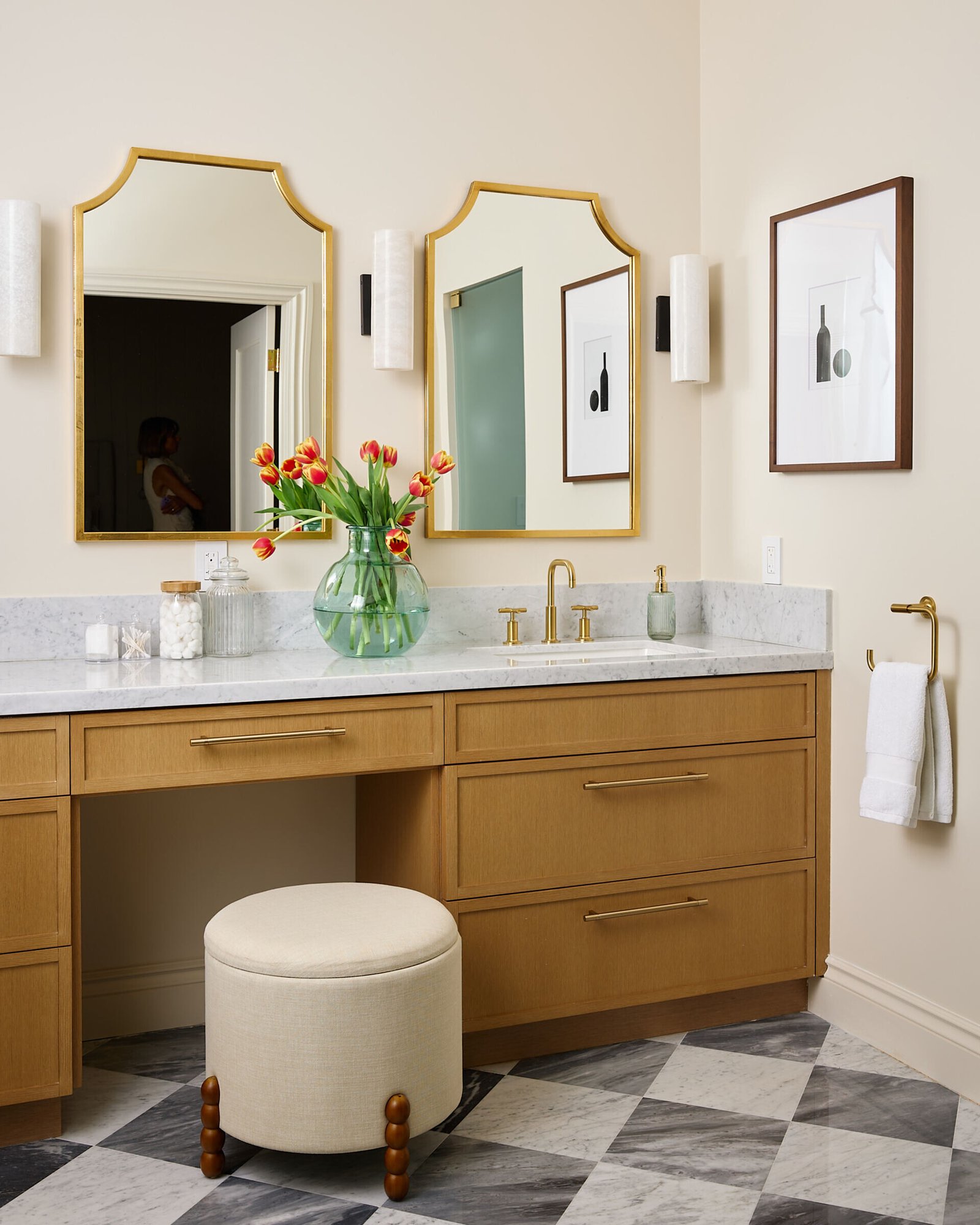 In this primary bath, quiet indulgence is the theme. The checkerboard marble floor adds a timeless graphic punch, offset by creamy wall tiles that bring in softness. A freestanding tub basks in sunlight and lake views, while the custom oak vanity topped with honed marble strikes a clean yet classic tone. Brass fixtures, gold-rimmed mirrors, and alabaster sconces round out the palette in glowing harmony.