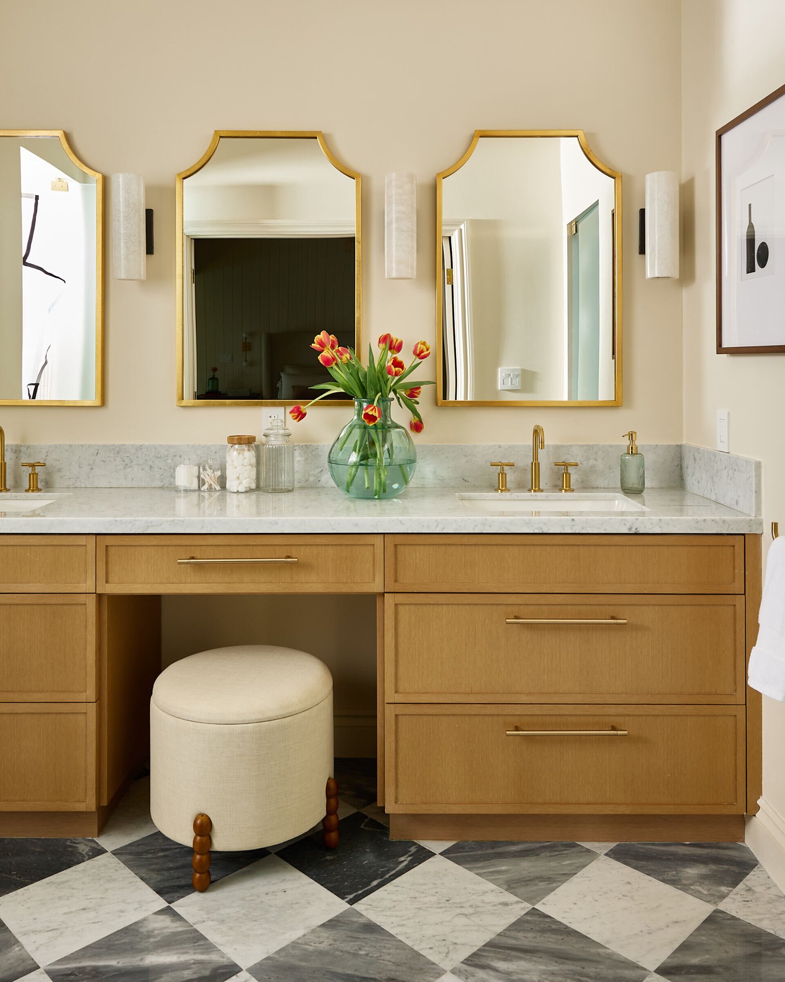 In this primary bath, quiet indulgence is the theme. The checkerboard marble floor adds a timeless graphic punch, offset by creamy wall tiles that bring in softness. A freestanding tub basks in sunlight and lake views, while the custom oak vanity topped with honed marble strikes a clean yet classic tone. Brass fixtures, gold-rimmed mirrors, and alabaster sconces round out the palette in glowing harmony.