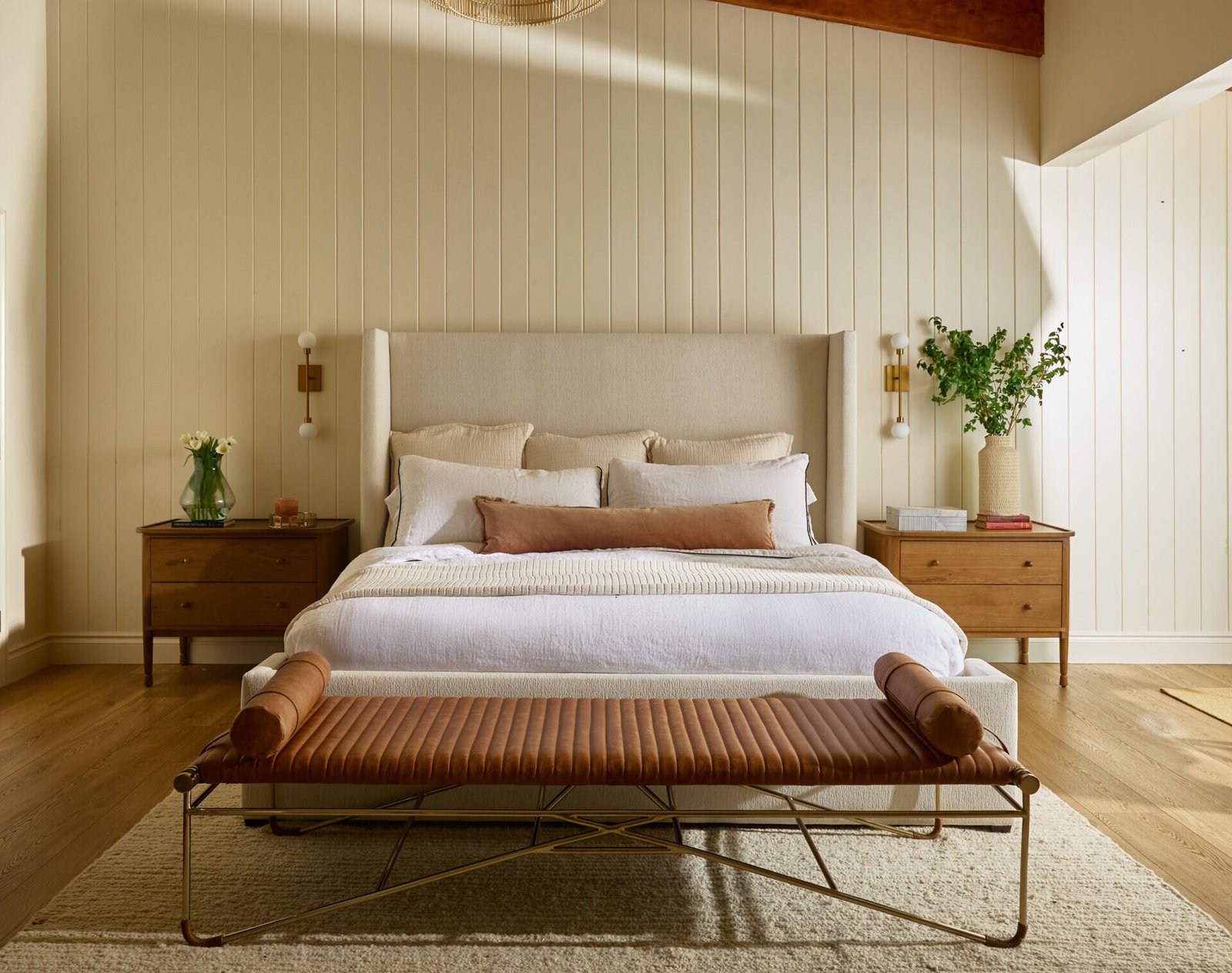 This primary bedroom has a tall upholstered wingback bed frame anchors the room, backed by light-colored vertical tongue-and-groove paneling that adds just enough architectural interest. Vaulted ceilings with exposed beams lift the space while keeping it grounded in natural warmth. A seating area by the windows is made for lake-gazing and late-night reads, and is framed by soft drapery.