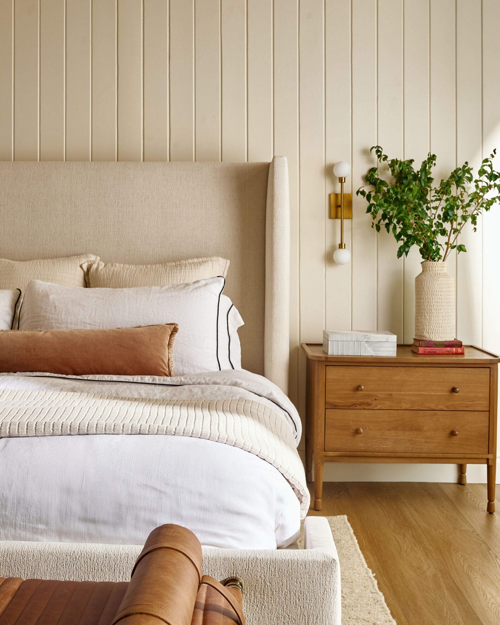 This primary bedroom has a tall upholstered wingback bed frame anchors the room, backed by light-colored vertical tongue-and-groove paneling that adds just enough architectural interest. Vaulted ceilings with exposed beams lift the space while keeping it grounded in natural warmth. A seating area by the windows is made for lake-gazing and late-night reads, and is framed by soft drapery.