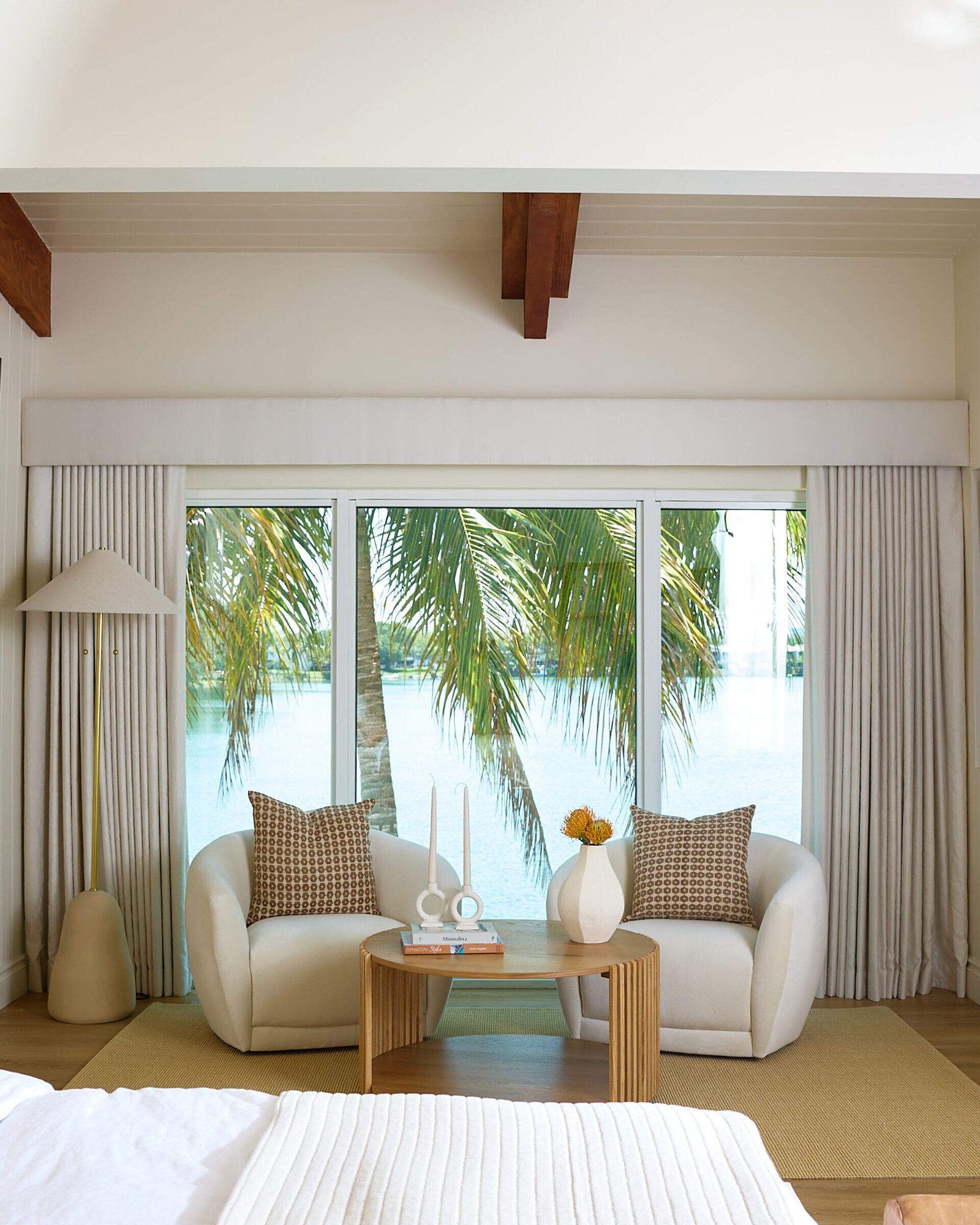 This primary bedroom has a tall upholstered wingback bed frame anchors the room, backed by light-colored vertical tongue-and-groove paneling that adds just enough architectural interest. Vaulted ceilings with exposed beams lift the space while keeping it grounded in natural warmth. A seating area by the windows is made for lake-gazing and late-night reads, and is framed by soft drapery.