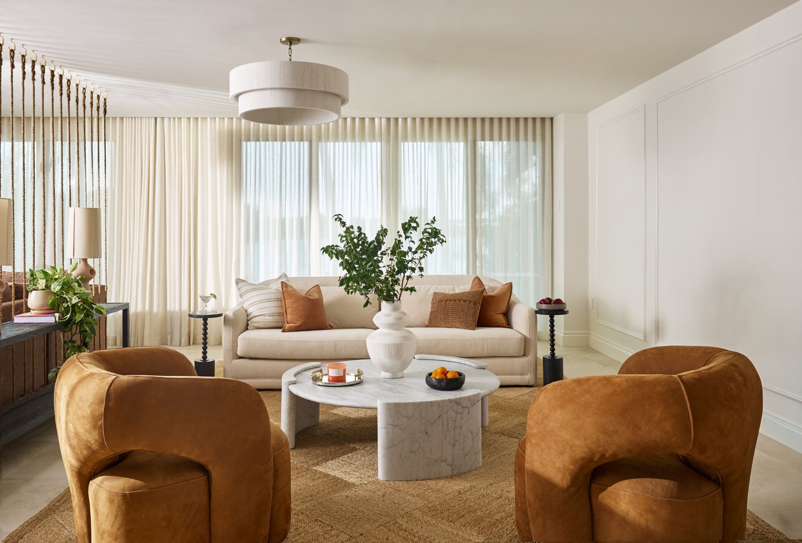 Warm neutrals and sculptural shapes make this more formal sitting room feel like a modern sanctuary. The soft cream sofa, styled with clay-toned pillows, sits against a wall of sheer drapes that gently filter daylight into the space. Two caramel velvet chairs add a rich, cozy texture, while the white marble coffee table introduces a cool, grounding contrast.