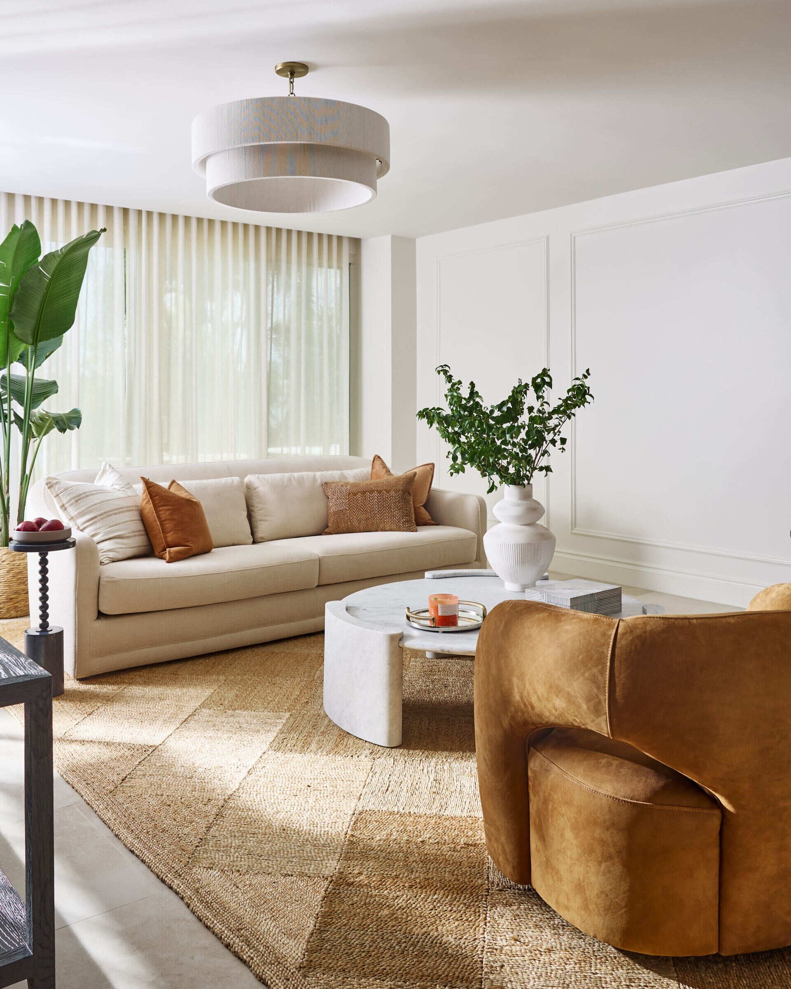 Warm neutrals and sculptural shapes make this more formal sitting room feel like a modern sanctuary. The soft cream sofa, styled with clay-toned pillows, sits against a wall of sheer drapes that gently filter daylight into the space. Two caramel velvet chairs add a rich, cozy texture, while the white marble coffee table introduces a cool, grounding contrast.