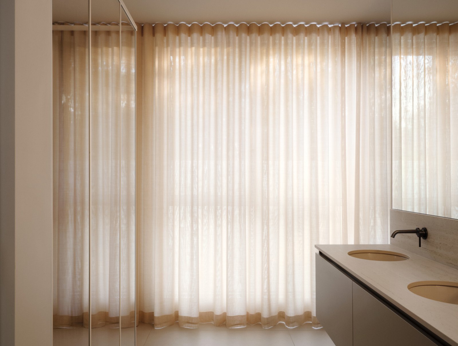 The primary bathroom is a quiet retreat, designed for comfort and calm. Sheer curtains filter soft light across a minimalist floating vanity with twin basins and a pale stone countertop. Muted metal fixtures add subtle contrast, while the mirrored above makes the room feel larger. With soothing tones, refined details, and no visual clutter, the ensuite feels spacious, tranquil, and perfectly suited for slow mornings. Two walk-in closets complete the suite with functional elegance.